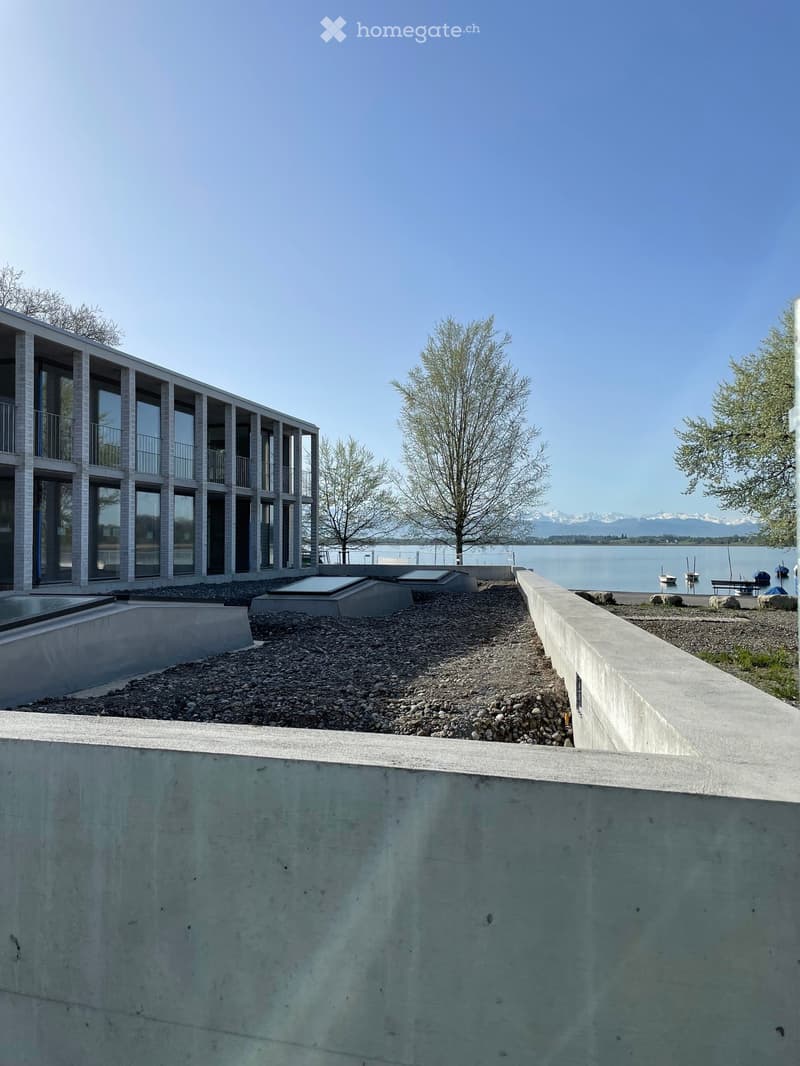 Modern building with balconies overlooking a lake and mountains, surrounded by gravel and trees.