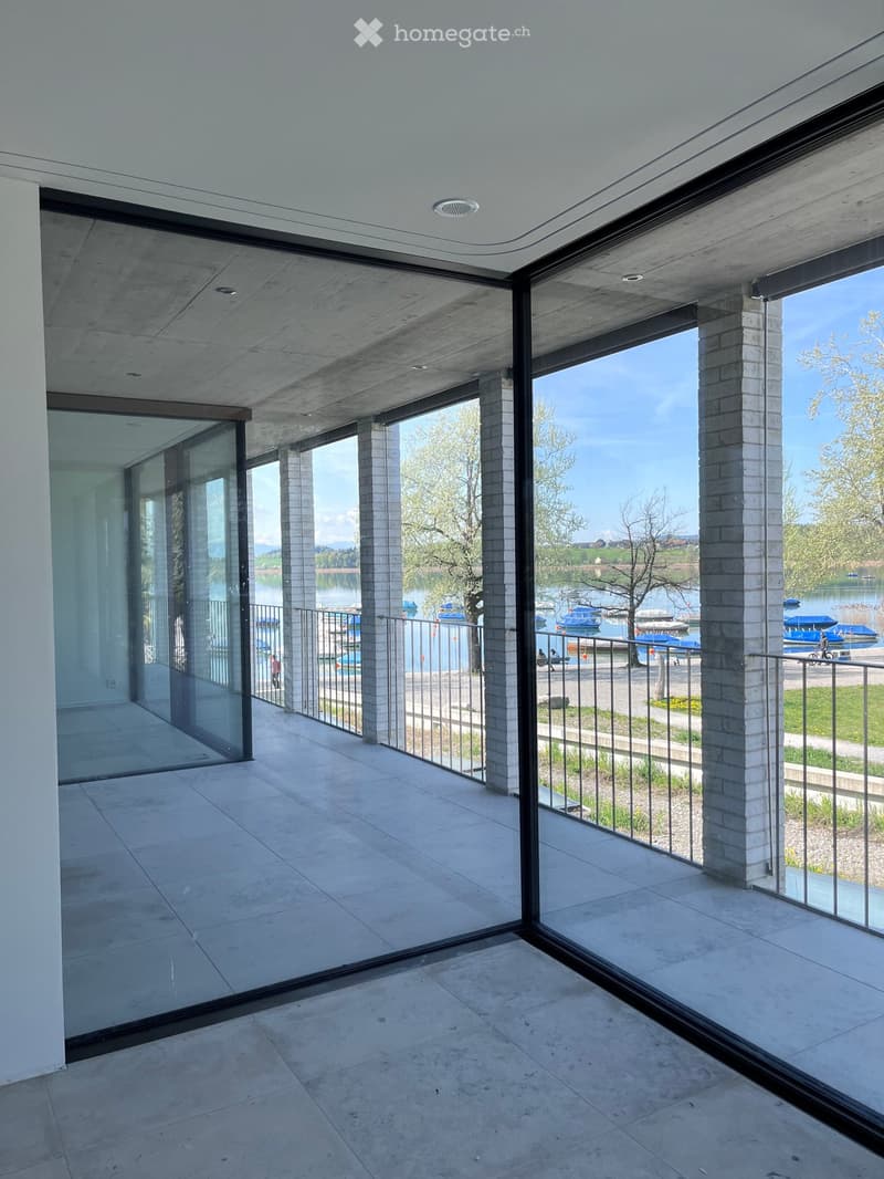 Modern balcony with large glass panels, concrete floor, and a view of a lake and boats.