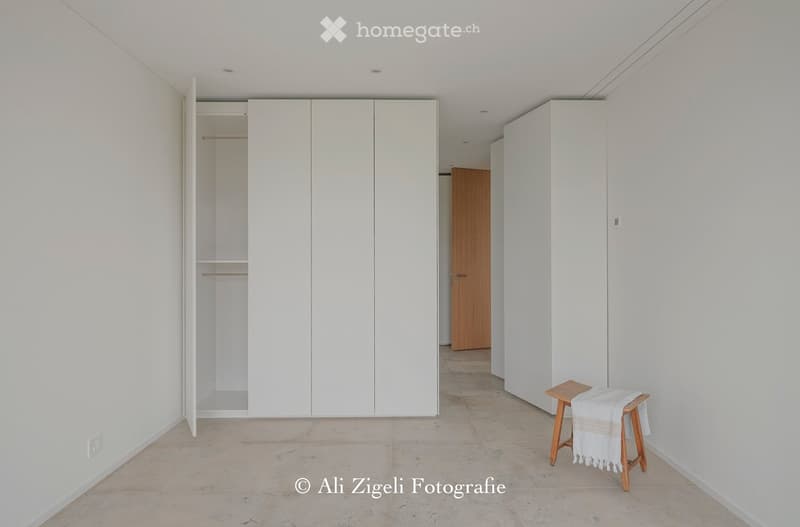 Minimalist room with white walls, tiled floor, built-in white wardrobes, and a small wooden stool with a white blanket.