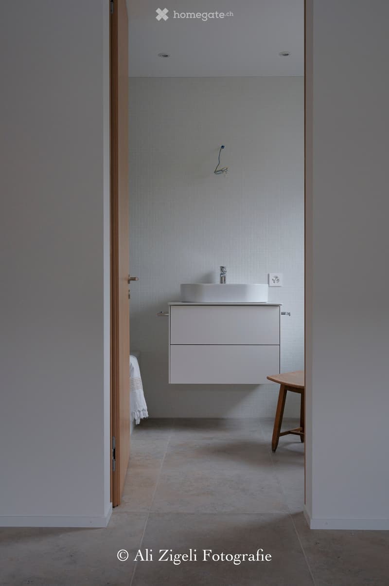 Modern bathroom with a white sink, cabinet, and wooden stool. Light grey tiled walls and a concrete floor.