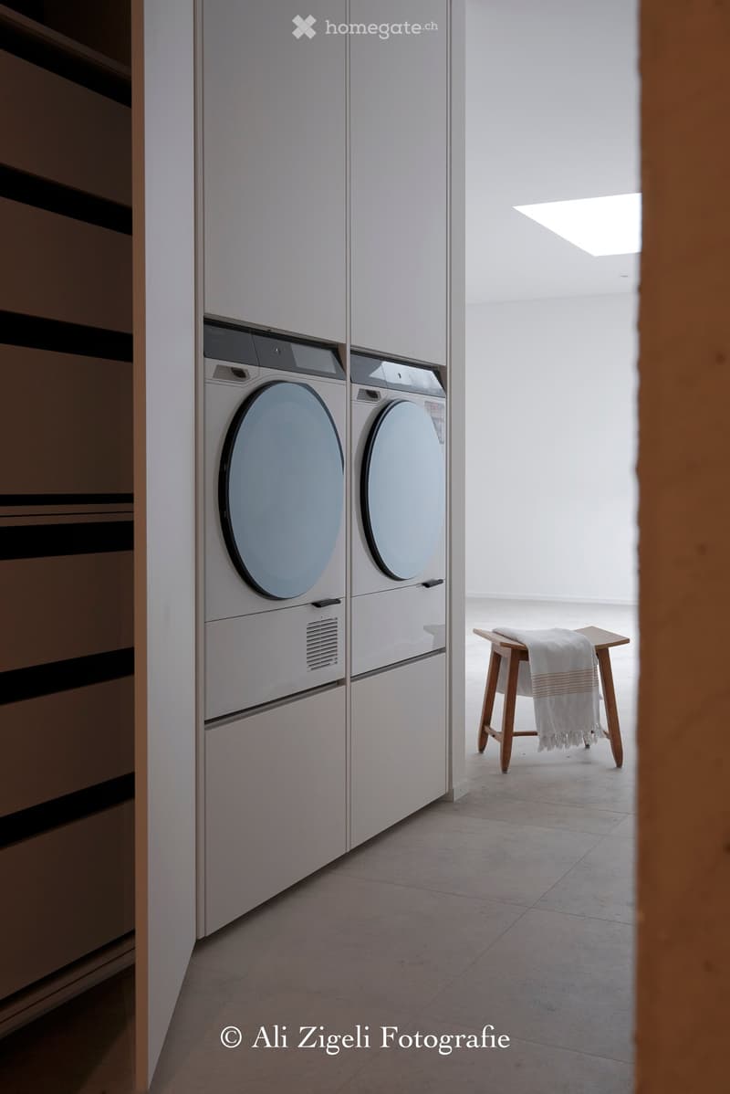 A modern, minimalist laundry area with a built-in washing machine and dryer unit, a small wooden stool with a folded towel, and a bright, airy space with natural light from a skylight.