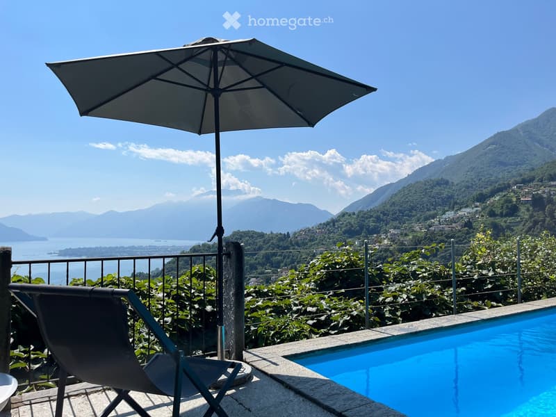 Outdoor area with a swimming pool, a lounge chair, and an umbrella, overlooking a scenic lake and mountain landscape.