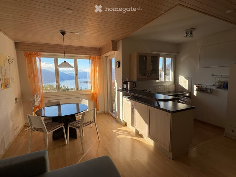 Open-plan kitchen and dining area with wooden flooring, white cabinets, black countertops, and a round dining table with four chairs. Large windows offer a scenic view of a lake and mountains.