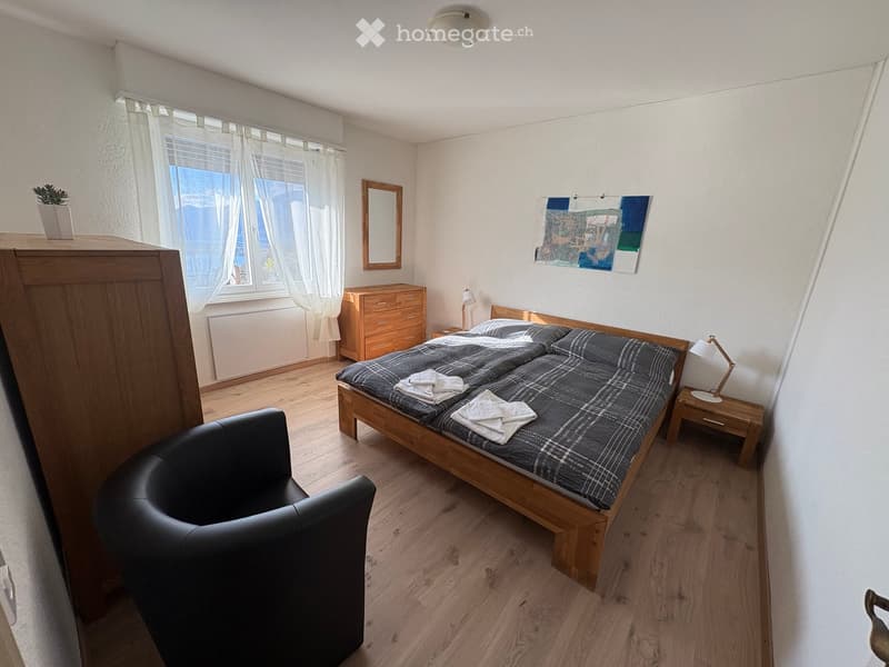 Cozy bedroom with a wooden bed, plaid bedding, a black leather armchair, a nightstand with a lamp, a dresser, a mirror, and a small plant on a cabinet.