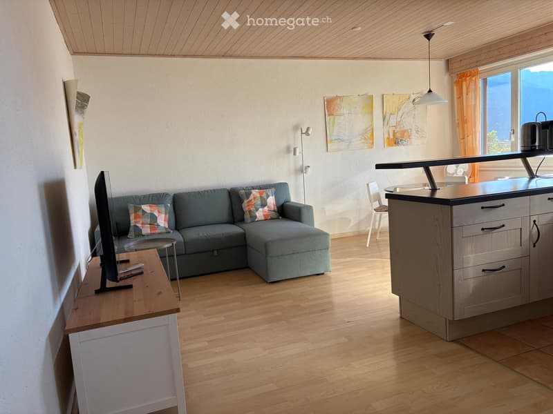 Modern living room with a grey sectional sofa, coffee table, TV stand, kitchen island, and artwork on the walls. The room features wooden flooring and large windows with orange curtains.
