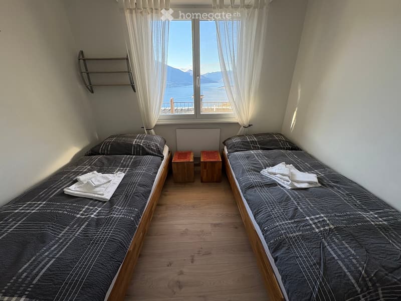 Cozy bedroom with two single beds, nightstands, and a scenic view of a lake and mountains through large windows.