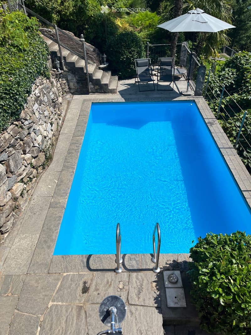 A rectangular swimming pool with clear blue water, surrounded by stone tiles, with two lounge chairs and an umbrella nearby, and lush greenery in the background.