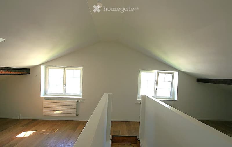 Empty attic room with wooden flooring, white walls, and two windows allowing natural light. The room features exposed wooden beams and a simple white railing along the edge of the loft area.