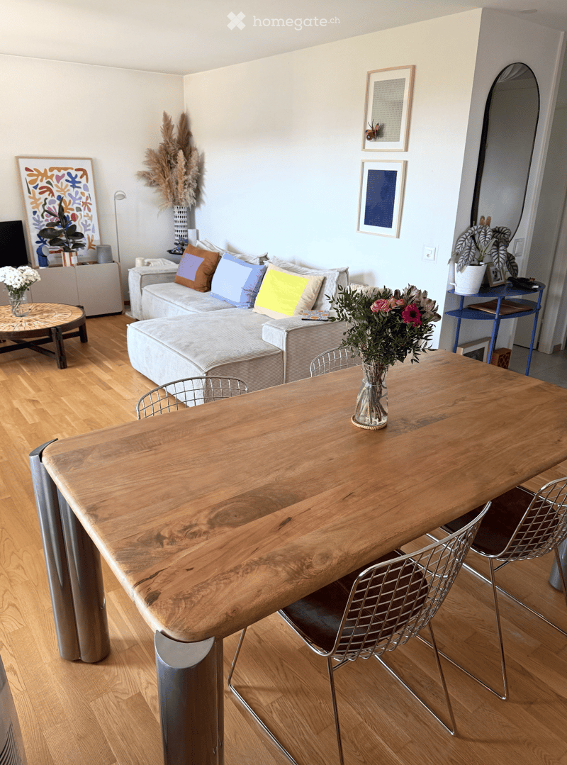 Modern living room with light wooden flooring, a large wooden table with metal chairs, a grey sofa with colorful pillows, a coffee table with a vase of flowers, and decorative artwork on the walls.