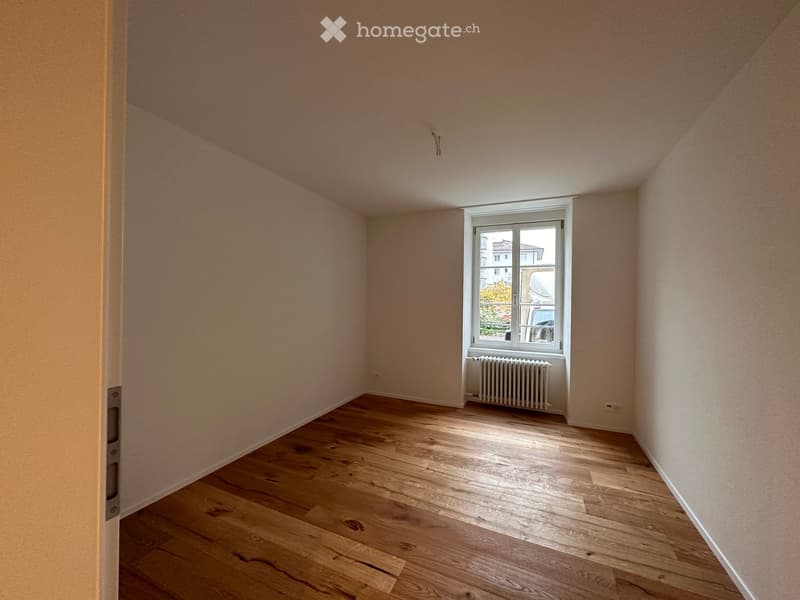 Empty room with wooden flooring, light-colored walls, and a single window with a radiator below it.