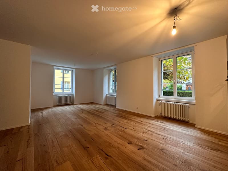 Empty room with wooden flooring, white walls, and three windows with radiators beneath them. Natural light streams in from the windows, and a single hanging light fixture is visible on the ceiling.