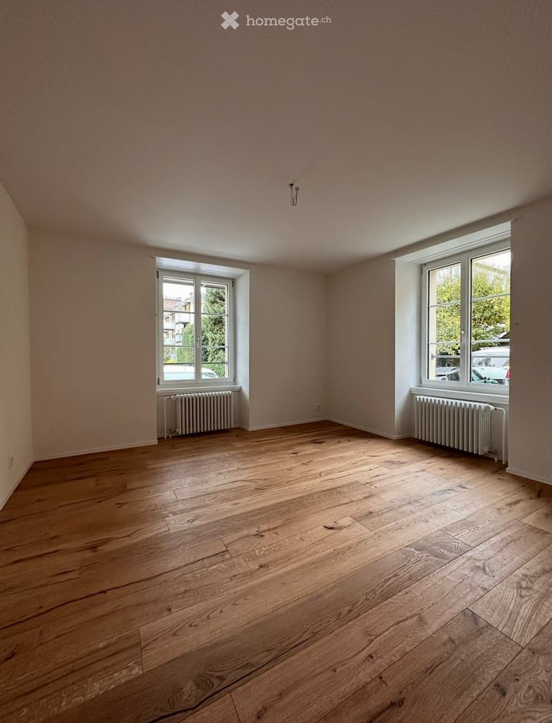 Empty room with wooden flooring, two windows, and radiators beneath them.