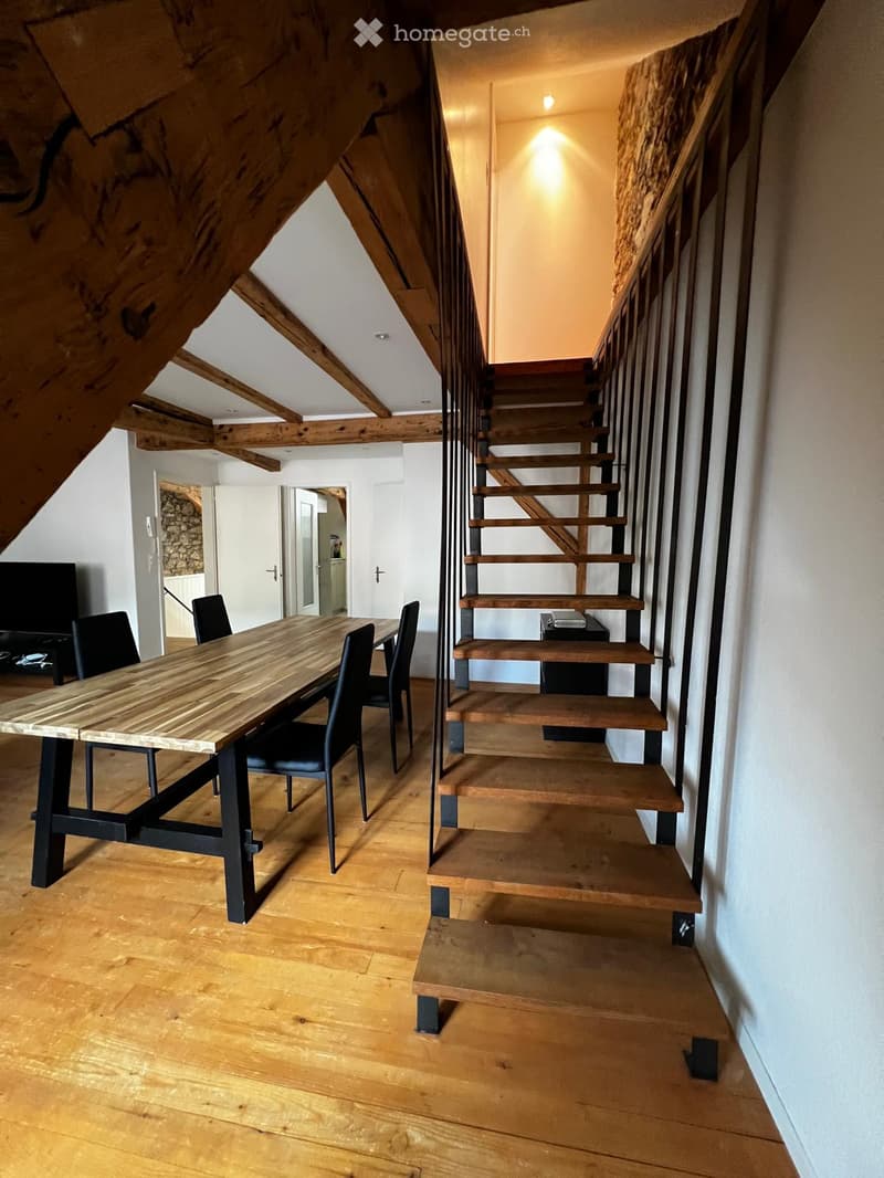 A modern, open-plan living and dining area with a wooden staircase leading to an upper level. The space features a large wooden dining table with black metal legs and matching chairs, a sleek black TV stand, and exposed wooden beams on the ceiling.