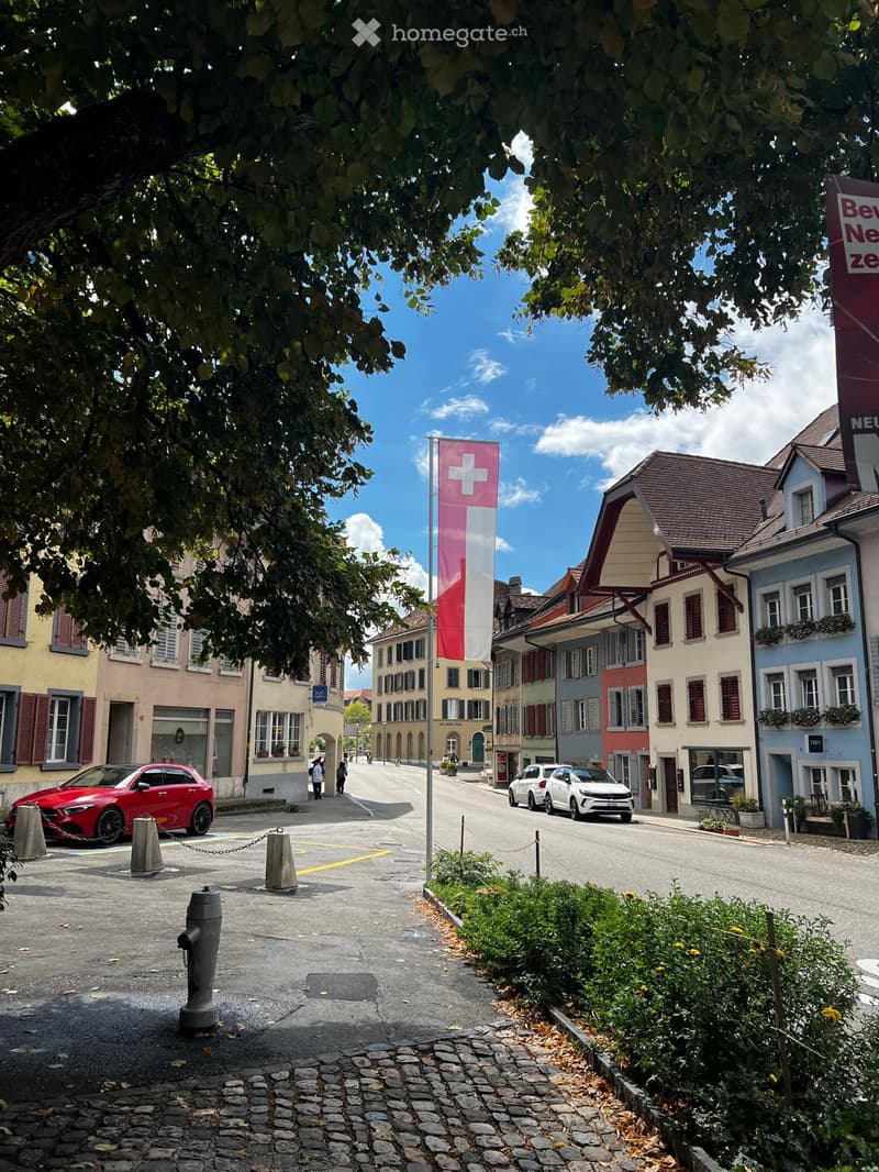 A picturesque European town street lined with colorful buildings, featuring a Swiss flag, parked cars, and pedestrians.