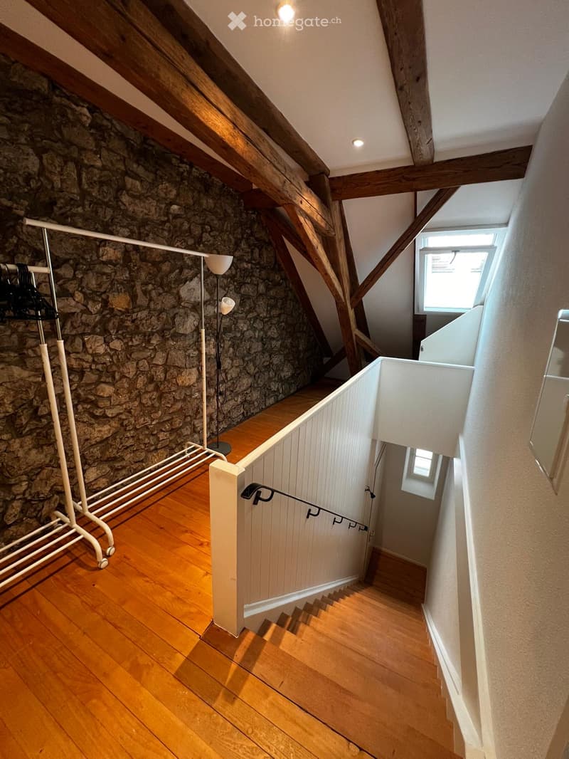 Wooden staircase with a white railing, stone wall, wooden floor, and a clothing rack with a floor lamp nearby.