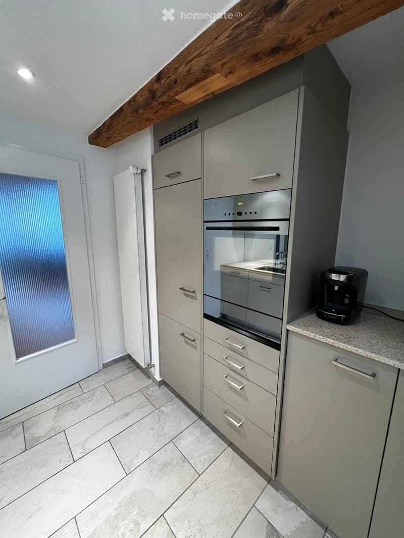 Modern kitchen with light grey cabinets, stainless steel oven, refrigerator, and a coffee maker on the counter. The floor is tiled, and the ceiling has exposed wooden beams.