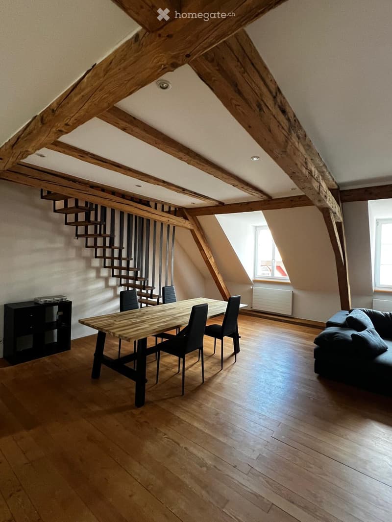 Open-plan living and dining area with exposed wooden beams, hardwood flooring, a sofa, a dining table with chairs, and a cabinet.