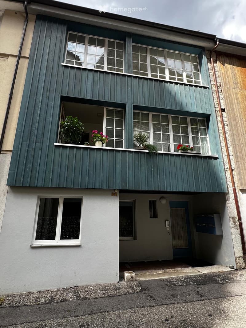 Two-story building with green wooden facade, large windows, and a small balcony with potted plants.
