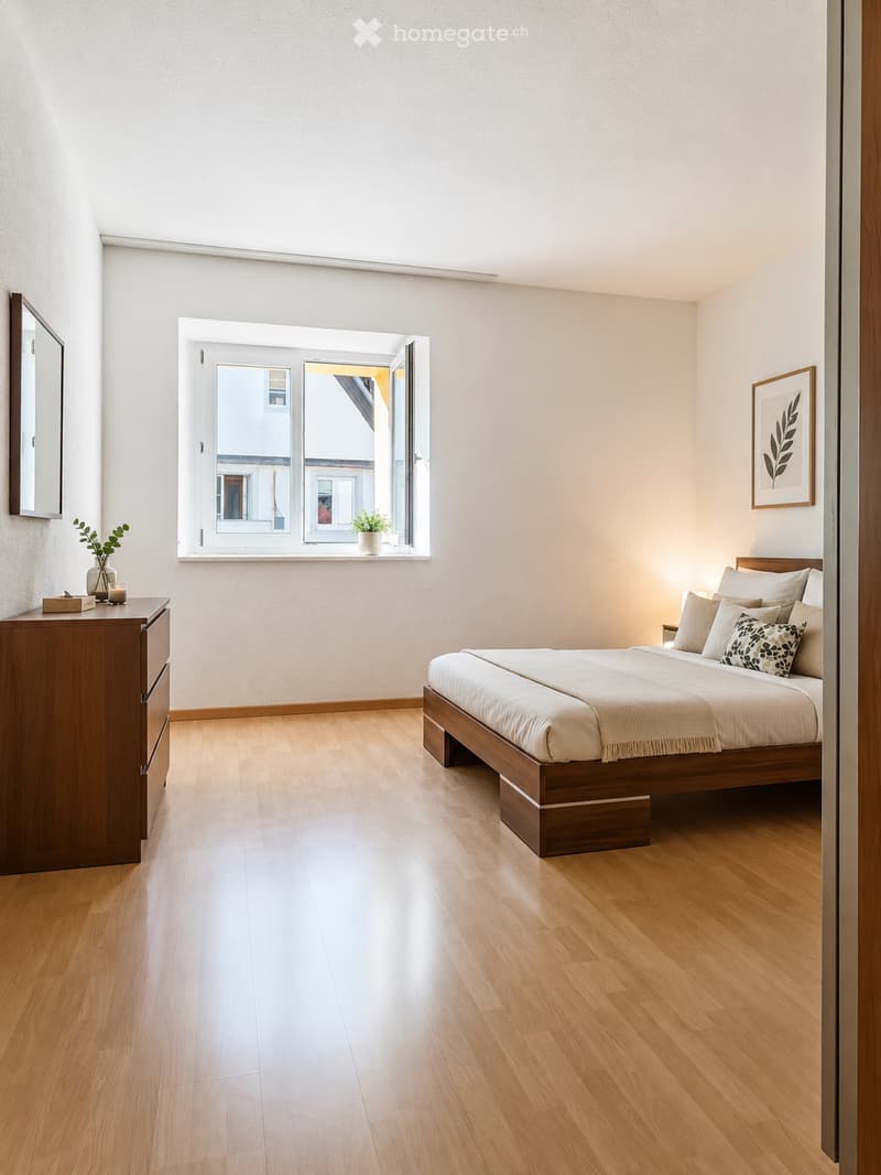Modern bedroom with wooden flooring, a large bed with multiple pillows, a nightstand with a lamp, a mirror, and a small table with a vase and candle.