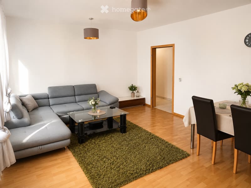 Modern living and dining area with a grey sectional sofa, glass coffee table, dining table with chairs, and decorative vases. The space features wooden flooring, a green rug, and pendant lighting.