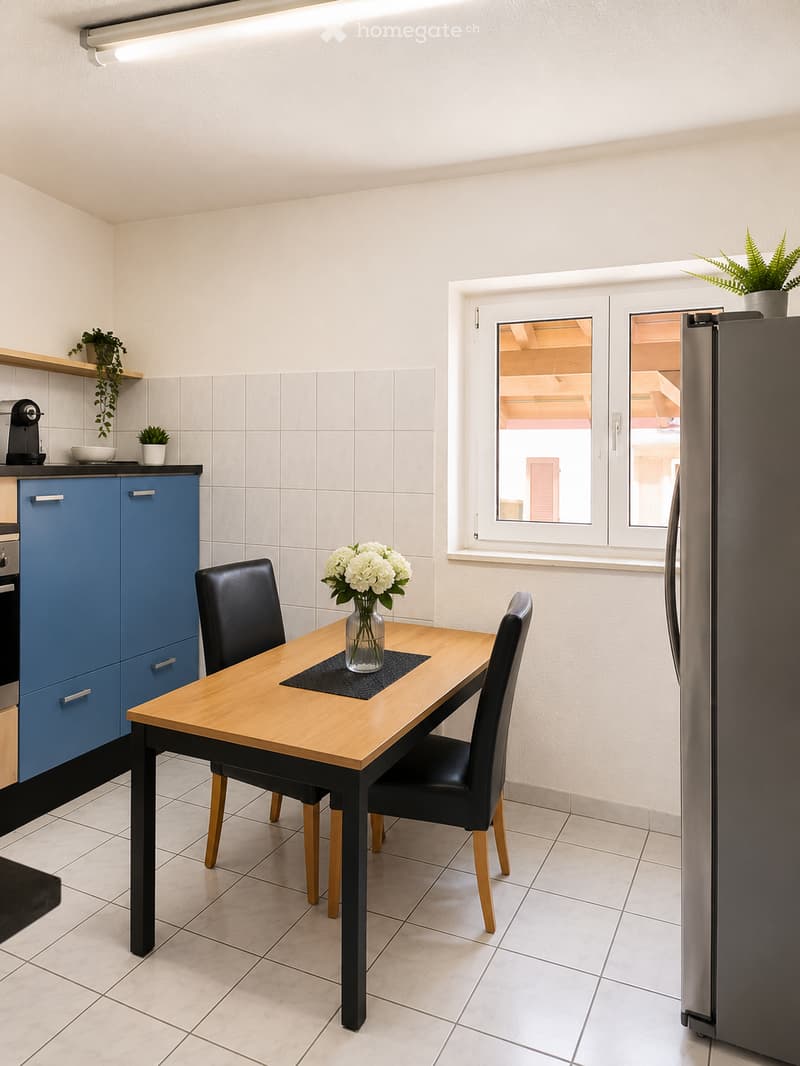 Modern kitchen with blue cabinets, a wooden dining table with two black chairs, a vase with white flowers, and a refrigerator. The room features tiled walls and a window with a view of a building.