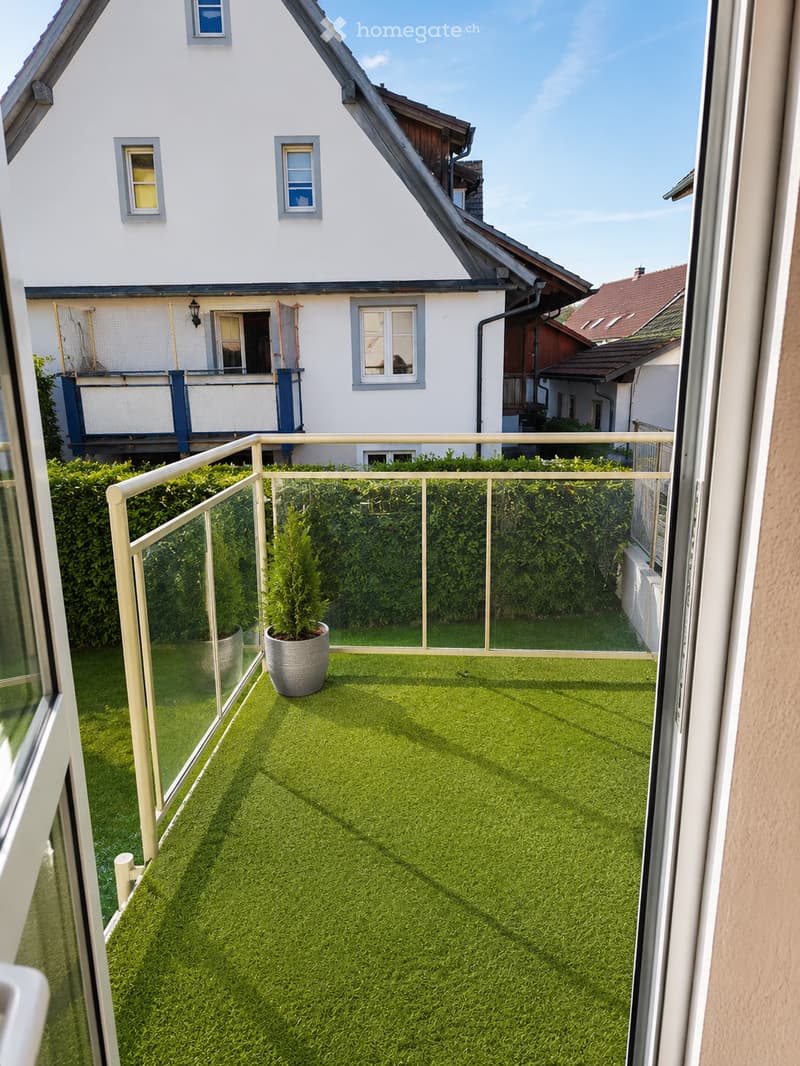 Balcony with artificial grass, metal railing, and potted plants, overlooking a garden and neighboring house.