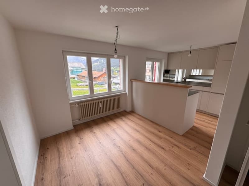 Bright, unfurnished room with wooden flooring, large windows offering a scenic view, and a small kitchen area with cabinets and a countertop.