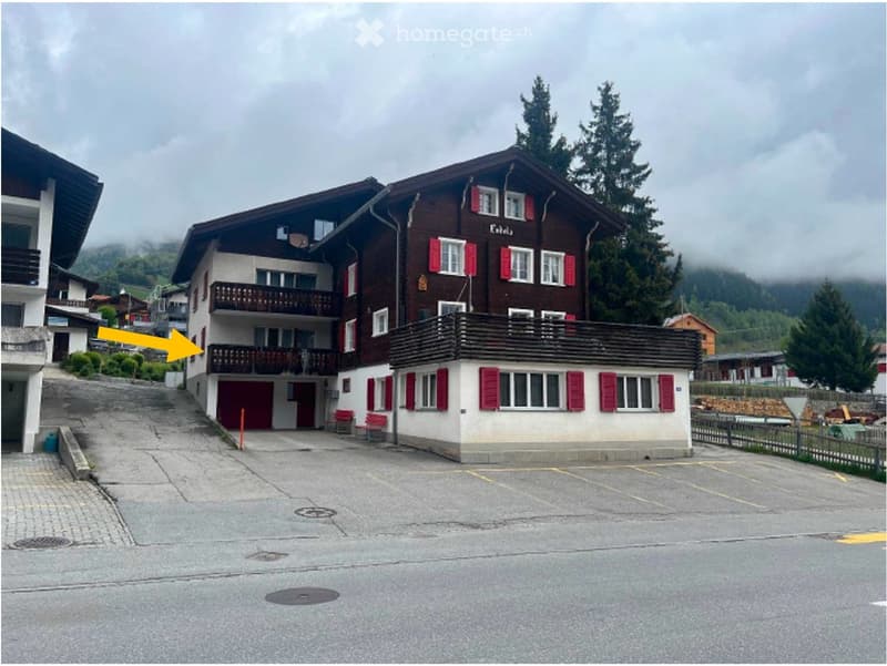 A two-story wooden building with red shutters and balconies, situated in a mountainous area with a paved parking area in front.