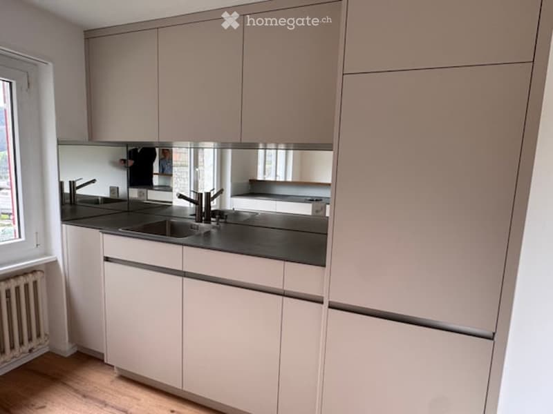 Modern kitchen with light-colored cabinets, a black countertop, a stainless steel sink, and a large mirror above the sink. The room features wooden flooring and a window on the left side.