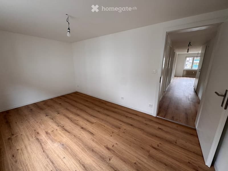 Empty room with wooden flooring and white walls, featuring a single light fixture and an open doorway leading to another room.