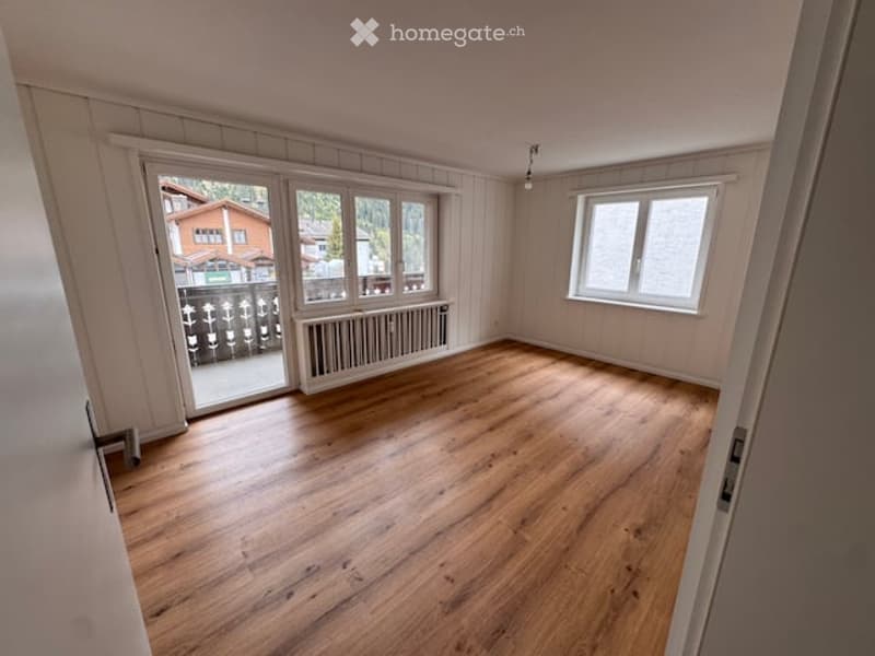 Empty room with wooden flooring, white paneled walls, large windows, and a sliding glass door leading to a balcony area.