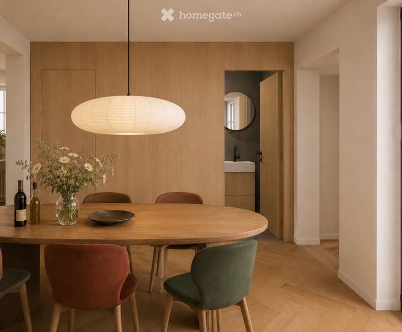 Modern dining area with a round wooden table, four chairs, a vase with flowers, two bottles, and a bowl on the table. A pendant light hangs above, and a doorway leads to a bathroom.