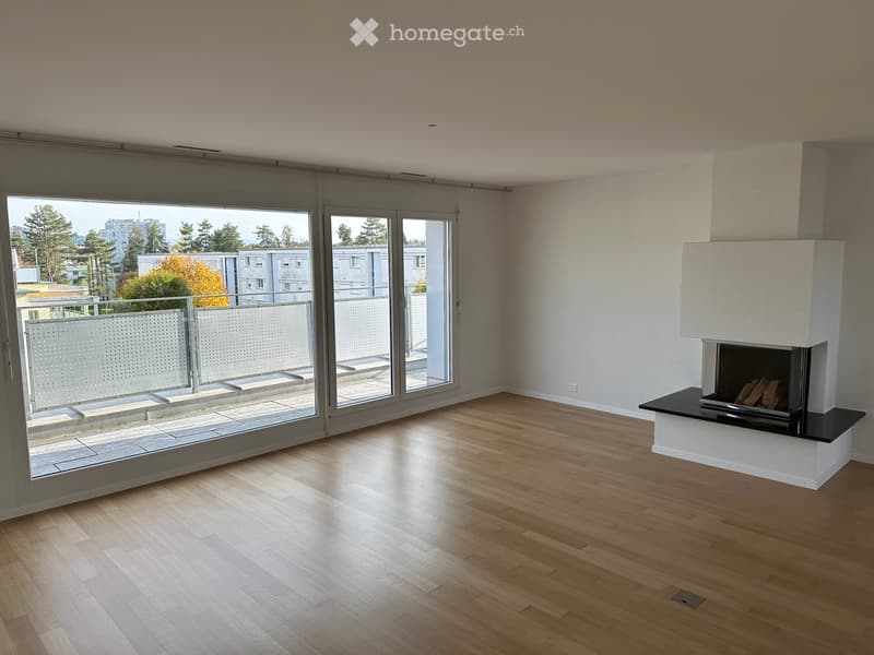 Bright, unfurnished room with wooden flooring, large windows, and a modern fireplace. The room features a balcony with a view of the city and surrounding greenery.