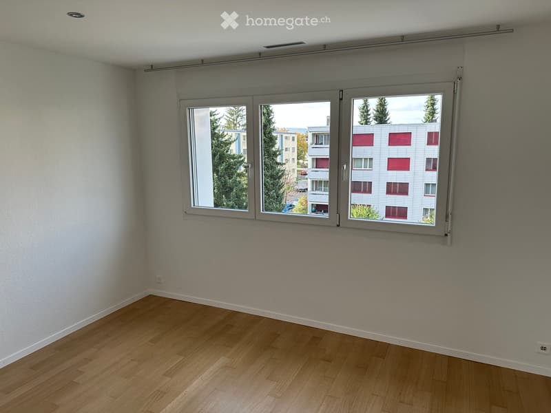 Empty room with wooden flooring and large windows showing a view of a building and trees outside.