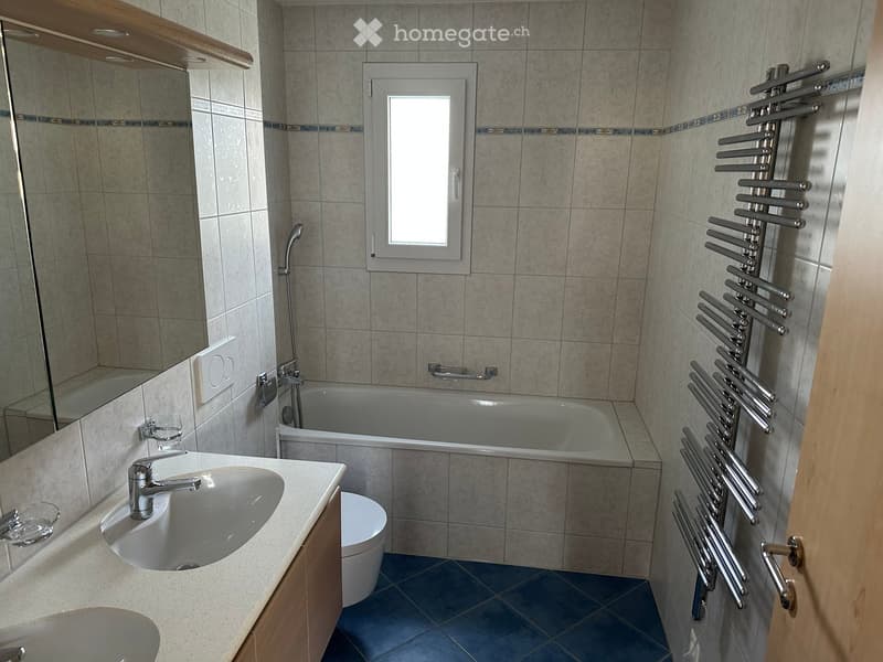 Modern bathroom with a white sink, bathtub, toilet, mirror, and a radiator on the wall. The room features tiled walls and a window for natural light.