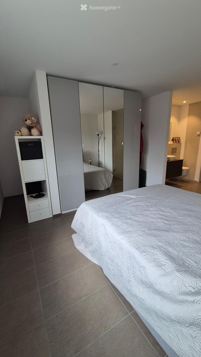 Modern bedroom with a large bed, mirrored wardrobe, small shelf with teddy bear, and an adjacent bathroom visible.