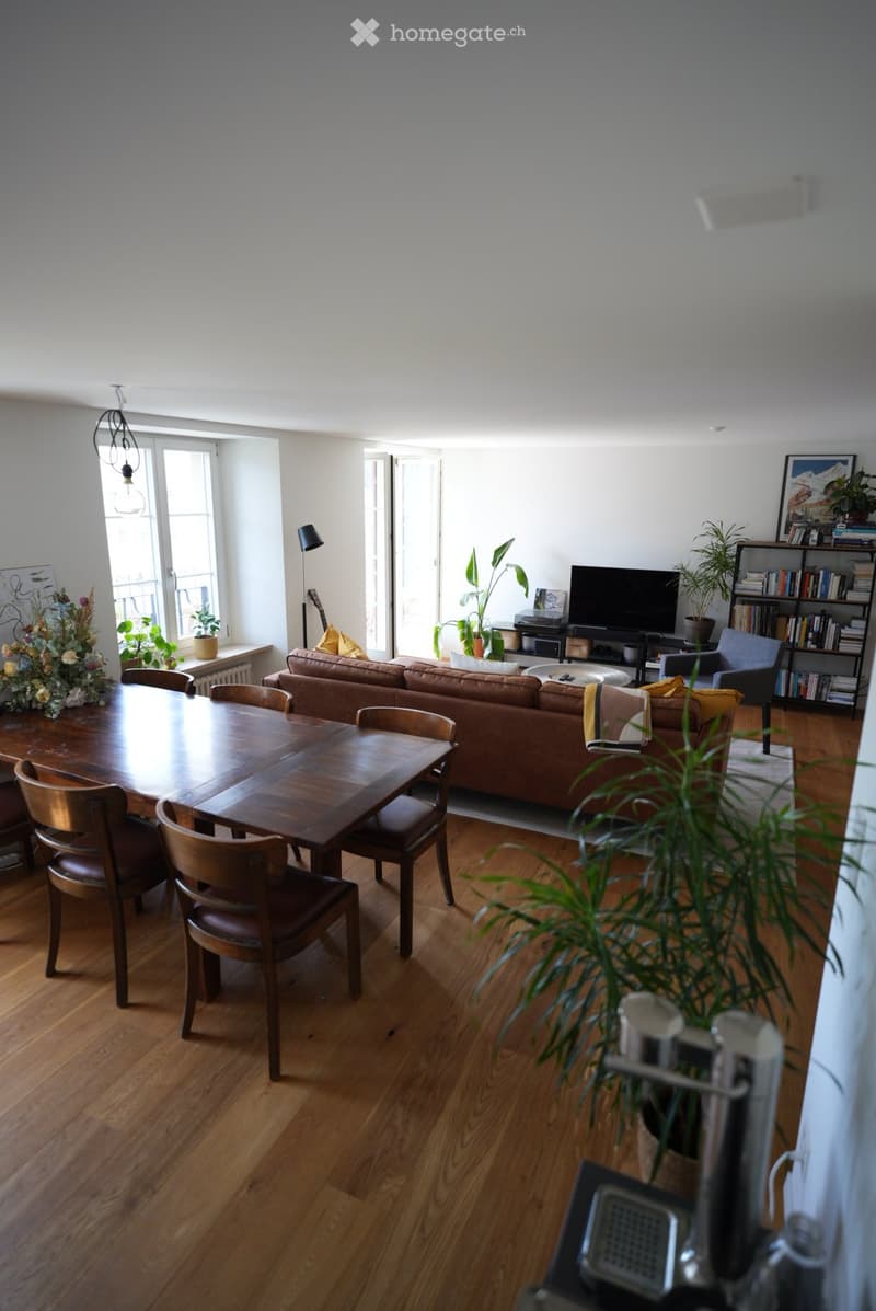 Bright living room with wooden flooring, a brown leather sofa, a wooden dining table with chairs, a TV, bookshelf, and decorative plants.