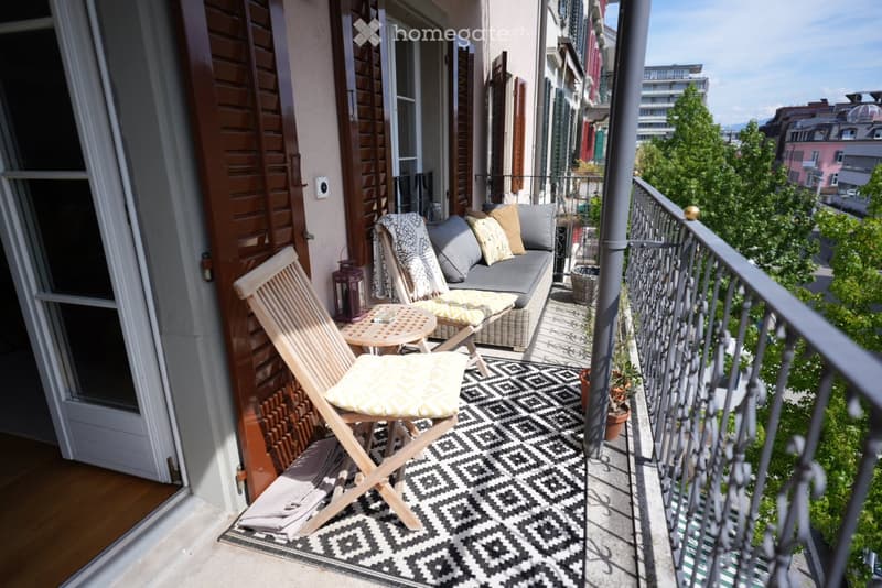 Balcony with a grey sofa, wooden folding chair, small table, patterned rug, and potted plants, overlooking a cityscape.