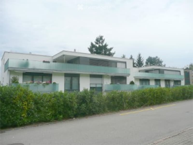 Modern multi-story residential building with balconies and large windows, surrounded by greenery.