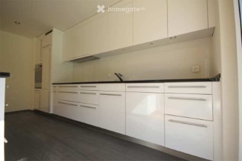 Modern kitchen with white cabinets, black countertop, and built-in oven. Dark wooden floor and sleek design.