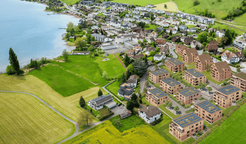 Aerial view of a residential area with multiple buildings, green spaces, and a lake in the background.