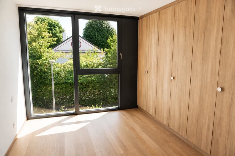 Empty room with light wooden flooring, white walls, and a large sliding glass door leading to a garden view.