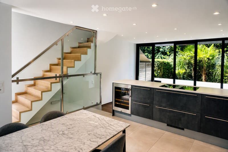 Modern open-plan space with a staircase leading to an upper level, a kitchen area with dark cabinets and stainless steel appliances, and a dining table with chairs.