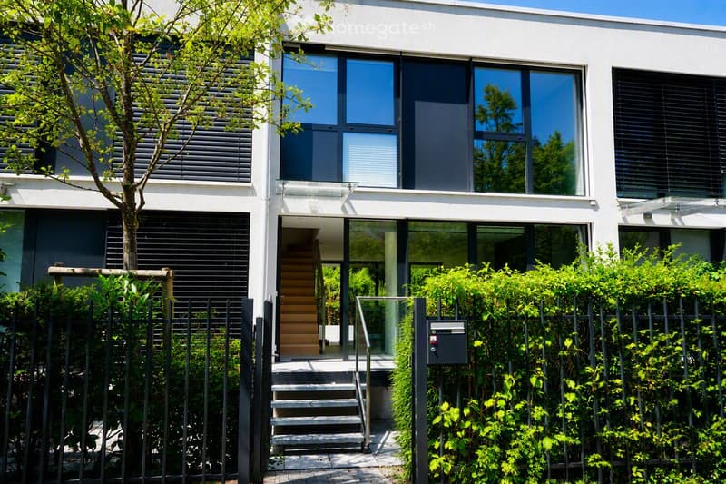 Modern two-story house with white exterior, large windows, and black shutters. Features a raised ground floor accessed by stairs, surrounded by a metal fence and greenery.