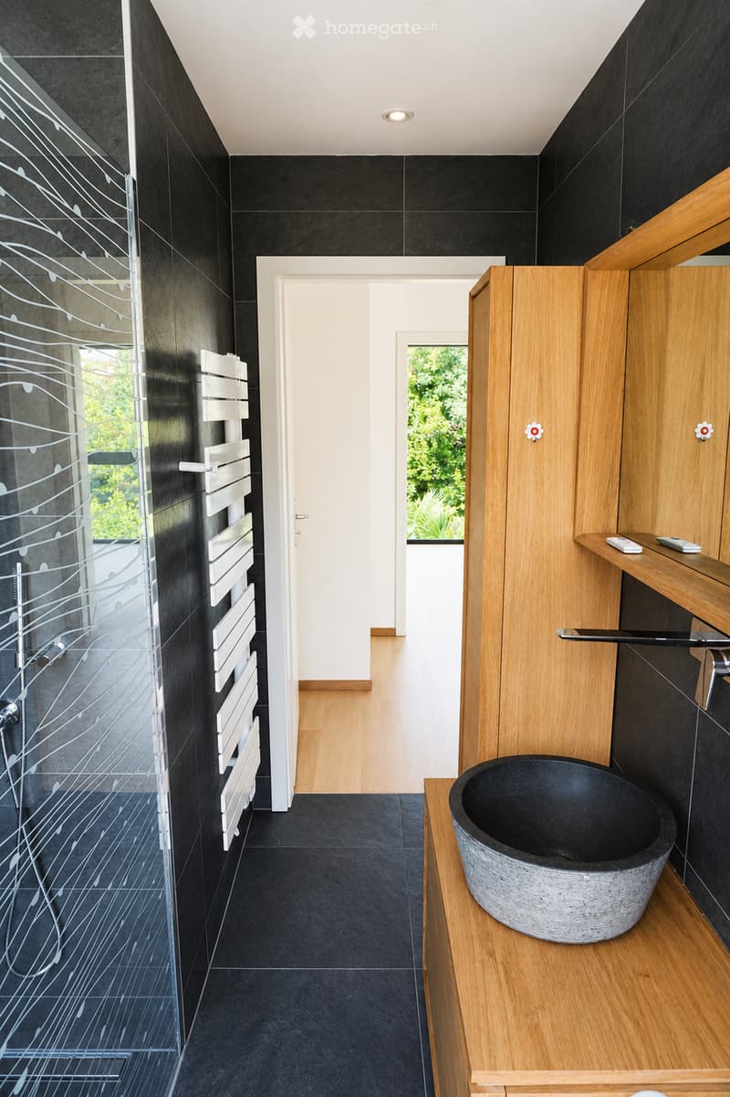 Modern bathroom with black tiles, a wooden shelf, a stone bathtub, and a glass shower enclosure with a radiator.