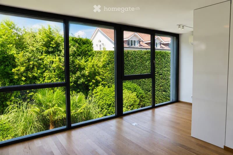Empty room with wooden flooring and large black-framed windows offering a view of greenery and neighboring houses.