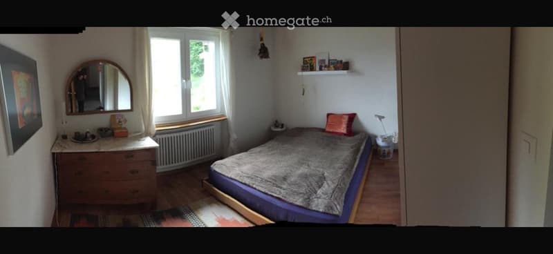 Cozy bedroom with a double bed, dresser, mirror, and shelves. The room features wooden flooring and large windows with white curtains.