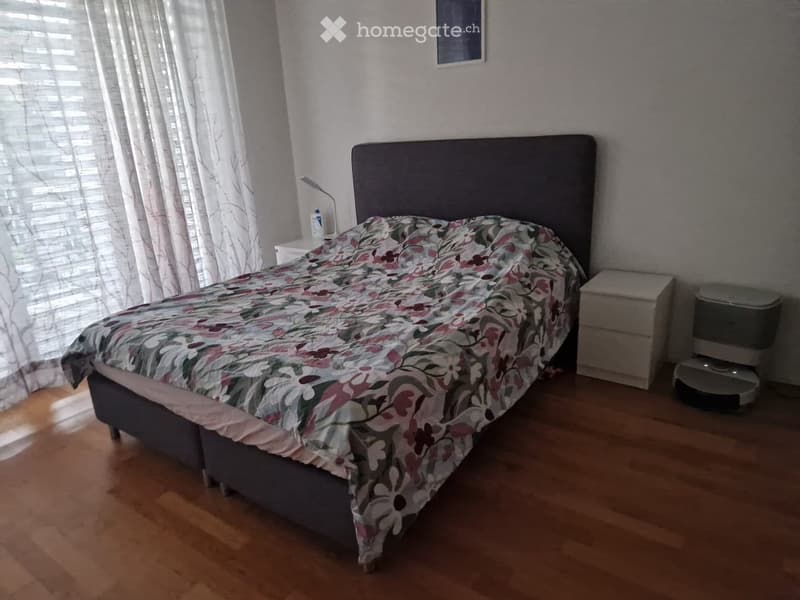 Bedroom with wooden flooring, a grey headboard bed with a floral duvet, a nightstand with a lamp, and a robot vacuum in the corner.