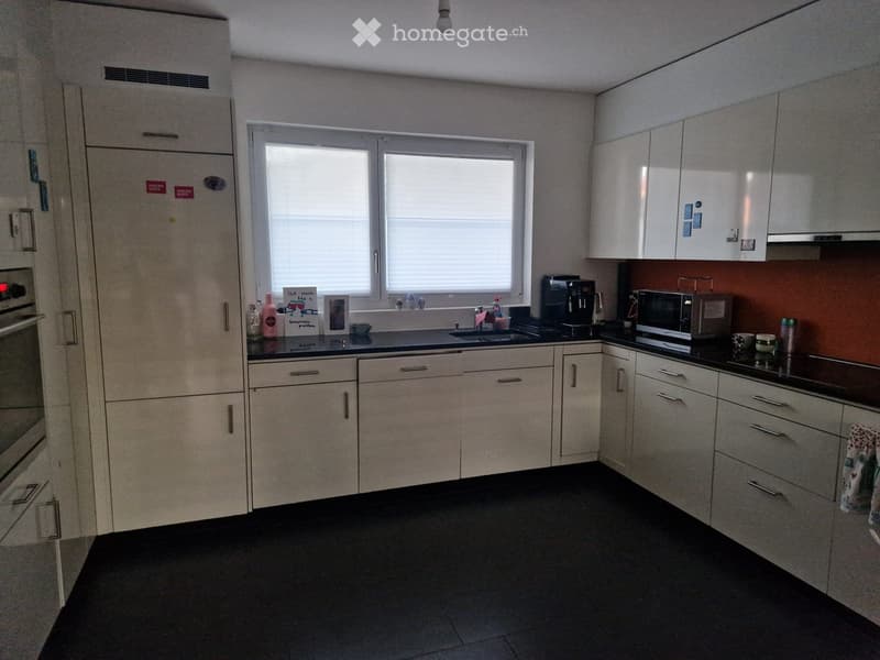 Modern kitchen with white cabinets, black countertops, and stainless steel appliances. Features a sink, coffee maker, microwave, and oven. The space is well-lit with natural light from a window.