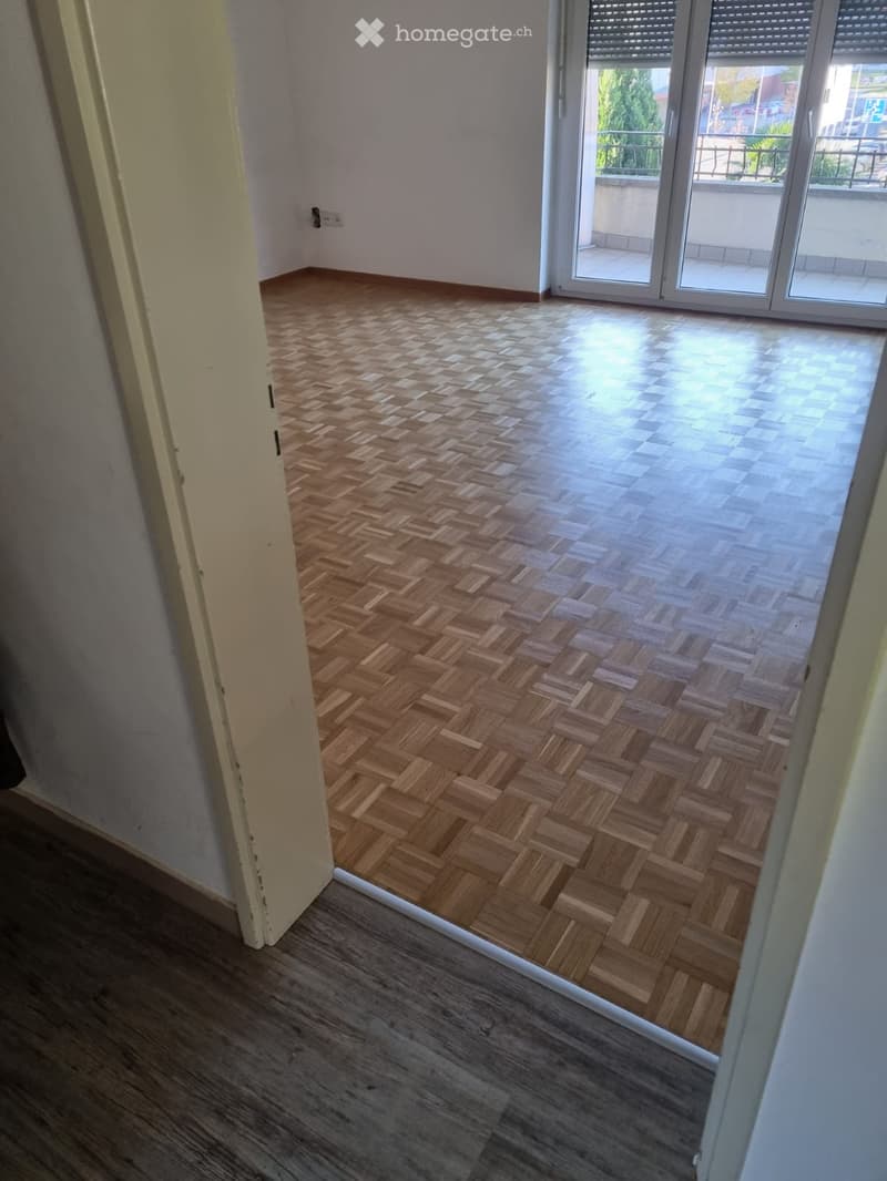 The image shows a room with a parquet floor. The floor is made of wooden planks arranged in a herringbone pattern. The room has large windows that provide natural light. The walls are painted in a light color.